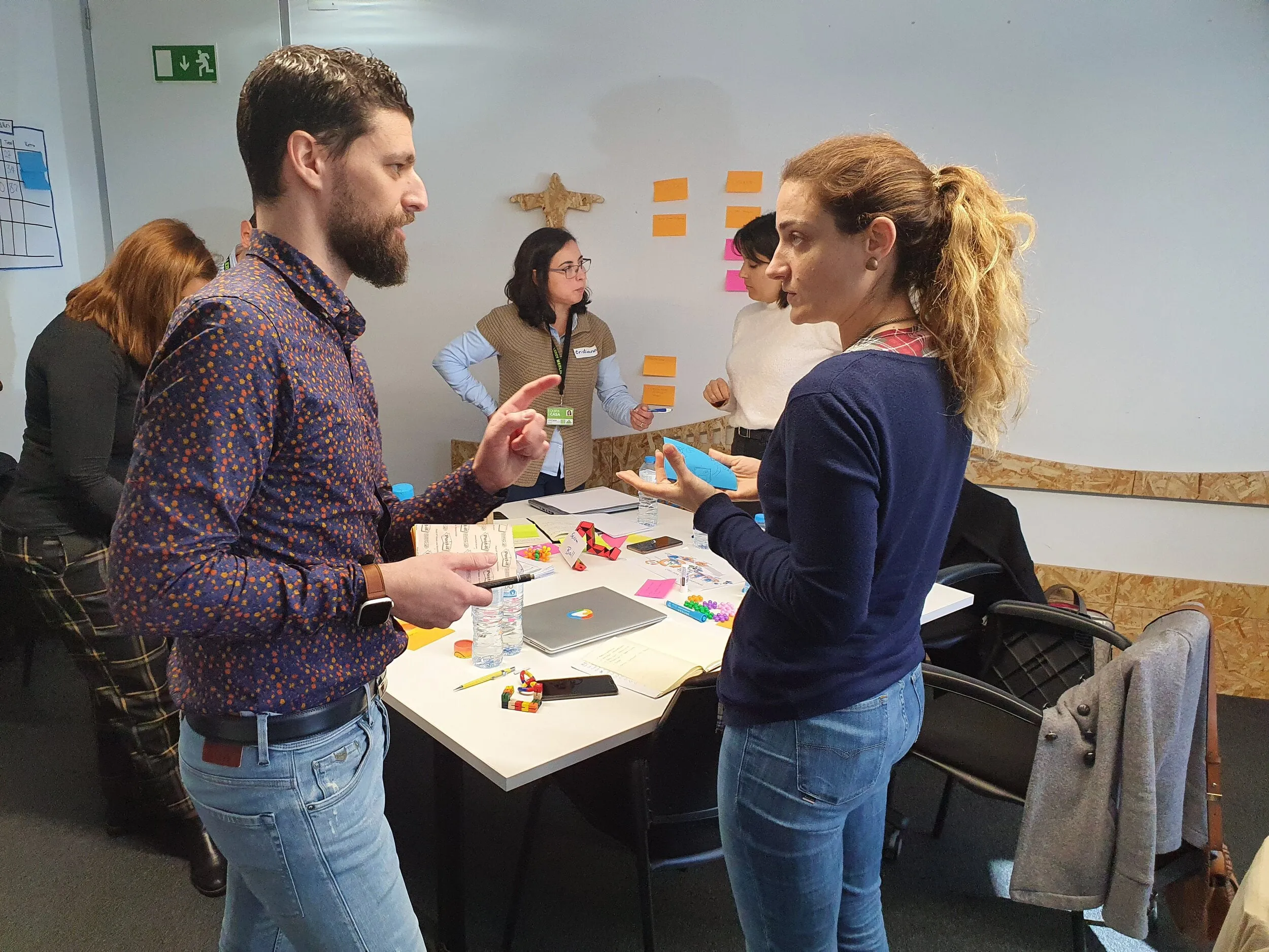 man in smart patterned shirt explaining something to a woman in an office, with other workers planning in the background