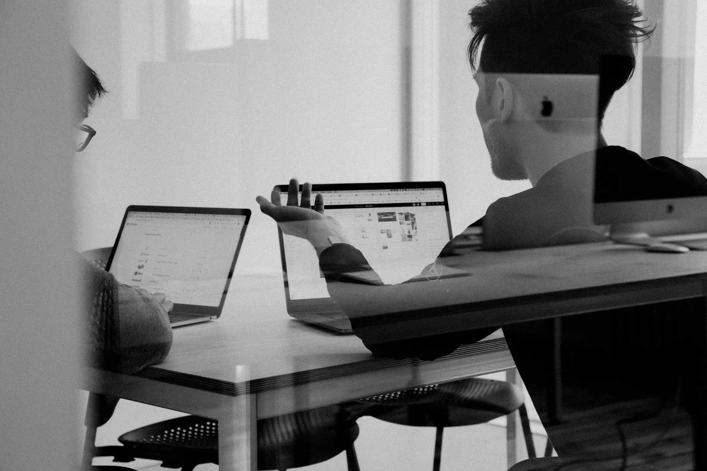 Two men in office behind glass wall talking and discussing topics with their laptops